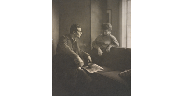 A black-and-white photograph of a White man and woman seated next to a sofa looking at one another.