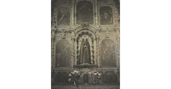 A black-and-white photograph of a sculpture of the Virgin Mary on an altar surrounded by paintings and decorative woodwork.