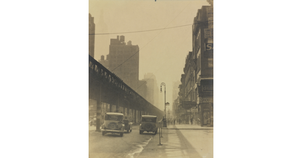 A sepia-toned photograph of vintage cars driving on a city street beside tall buildings and under an elevated train track.