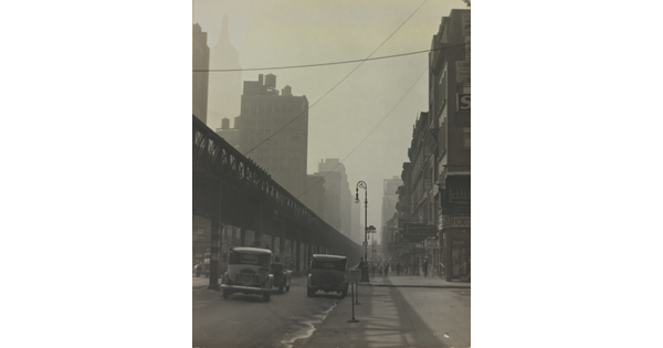 A black-and-white photograph of vintage cars driving on a city street beside tall buildings and under an elevated train track.