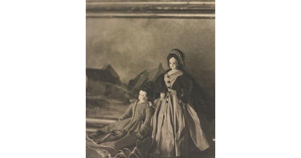 A black-and-white photograph of two light-colored dolls wearing formal 19th century European dresses posed in front of a landscape painting.