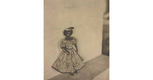 A black-and-white photograph of a dark-colored doll with curly hear wearing a patterned dress and hat posed against a wall.