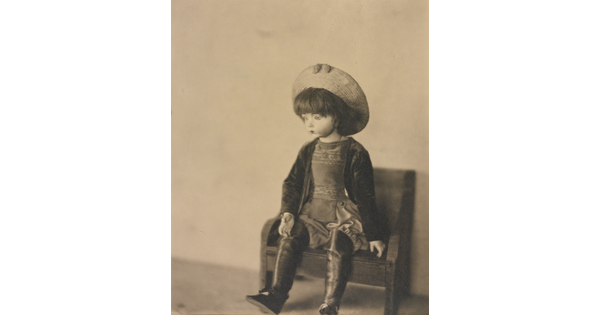 A sepia-toned photograph of a light-colored doll wearing a hat and boots sitting on a small bench.