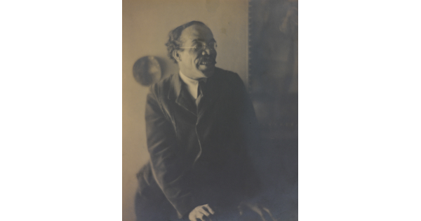 A sepia-toned, soft-focus photograph of a man wearing a suit and glasses leaning on something in front of him and looking away from the camera.