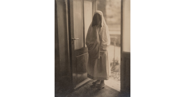 A black-and-white photograph of a woman in a doorway covered from head to toe in a white niqab with dark netting over her face.