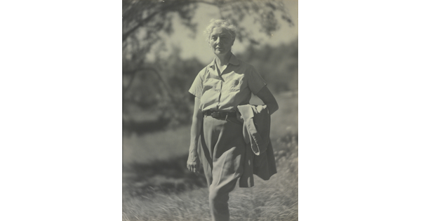 A black-and-white photograph of an older White woman with short, white hair standing outside holding a jacket in the crook of her arm.
