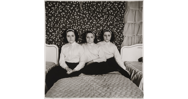 A black-and-white photograph of three teenage, triplet, girls dressed alike and seated on a twin bed.