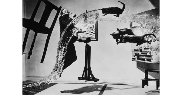A black-and-white photograph of a man painting on an easel along with a stream of water, a chair and three cats, all hovering in the air.