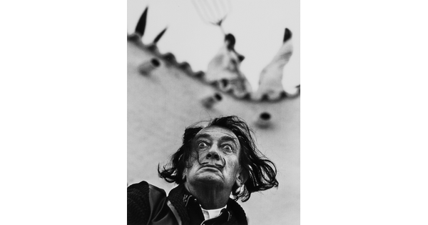 A black-and-white photograph looking up at a White man with medium-length wind-blown dark hair, a pencil mustache, wide eyes, and a frown.