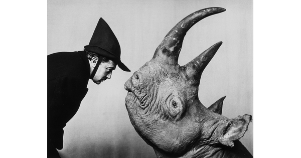 A black-and-white photograph of a White man wearing a Peter Pan-style hat facing a rhinoceros; both human and animal in profile.