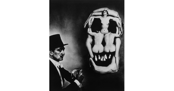 A black-and-white photograph of Salvador Dali in profile next to several nude women posed in the shape of a skull.