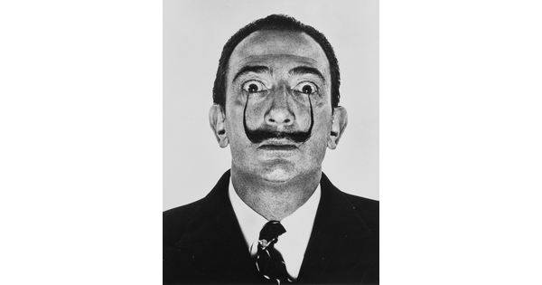 A black-and-white portrait photograph of a White man with a pencil mustache, the tips curved up all the way to his wide eyes.