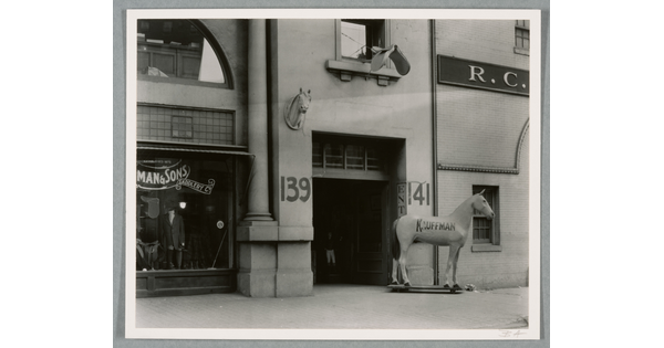 A black-and-white photograph of a storefront with a large statue of a horse, the word "Kauffman" painted on its side, next to the entrance.