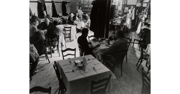 A black-and-white photograph of people sitting at multiple tables inside a restaurant.