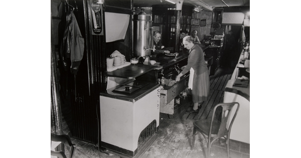 A black-and-white photograph of a woman working in a restaurant while people eat at a counter.
