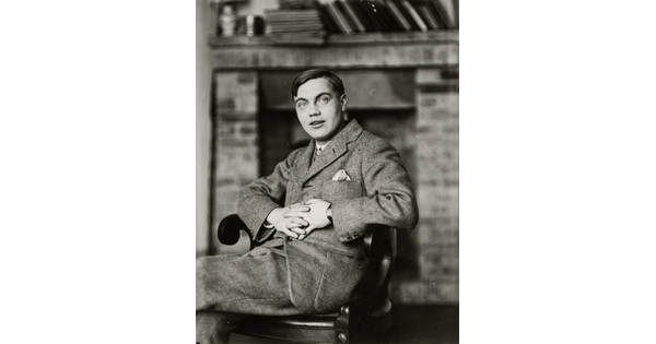 A black-and-white portrait photograph of a White man in a suit sitting in a wood chair with his hands clasped in front of him.