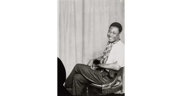 A black-and-white portrait of a Black man sitting with his legs crossed, smiling at the camera.