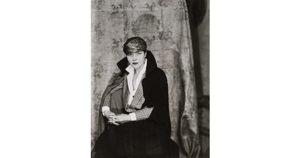 A black-and-white portrait of an elaborately dressed White woman seated in a chair with a coat draped over her shoulders.