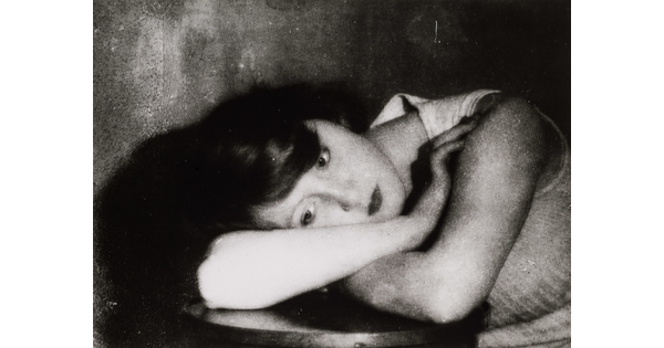 A black-and-white portrait photograph of a White woman leaning over a table and resting her head on her arms.
