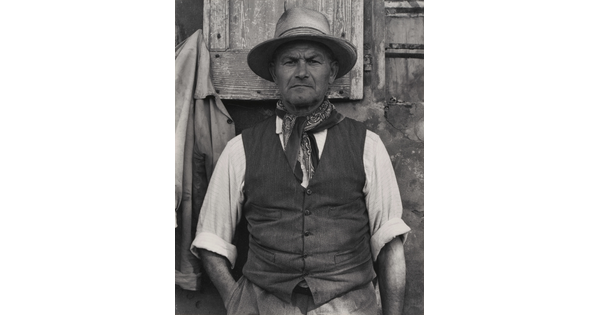 A black-and-white portrait photograph of a medium-skinned man from the waist up wearing a hat, bandanna tied around his neck, vest, and a shirt rolled to his elbows.