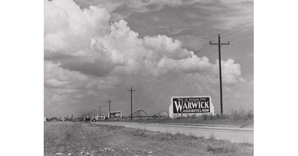 A black-and-white photograph of a rural road lined with billboards and powerlines, and a rollercoaster in the distance.