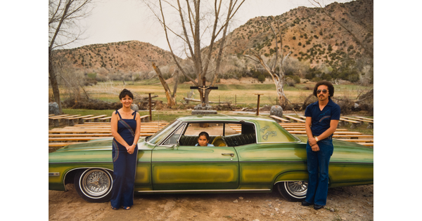 A color photograph of two adults standing in front of a green car that's in profile with a child sitting in the driver's seat.