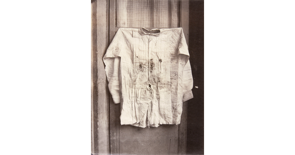 A black-and-white photograph of a shirt with bullet holes and bloodstains hanging on a door.