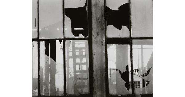 A black-and-white photograph of broken window panes in what looks like a factory building.