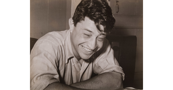 A black-and-white photograph of a seated White man laughing and leaning forward on his arms.