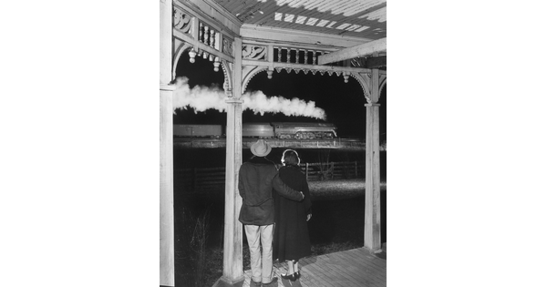 A black-and-white photograph of a couple standing under a covered porch watching a steam train pass by at night.