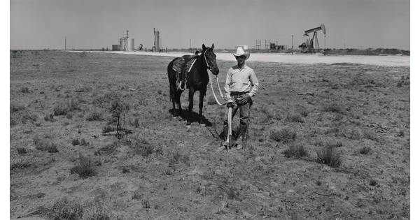 A black-and-white photograph of a man wearing sunglasses and a cowboy hat standing next to his horse, an oilfield and pumpjacks in the background.