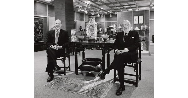 A black-and-white photograph of two smiling White men in suits sitting on either side of a table displaying Asian decor.