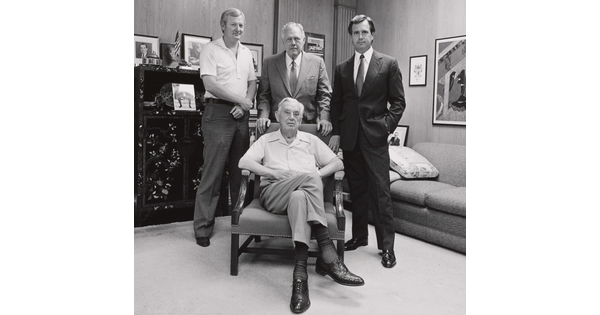 A black-and-white photograph of four White men of varying ages, three standing and one seated in an armchair, in an office.