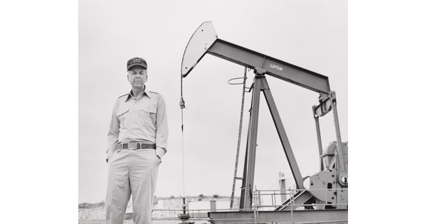 A black-and-white photograph of an older White man standing in front of a pump jack.