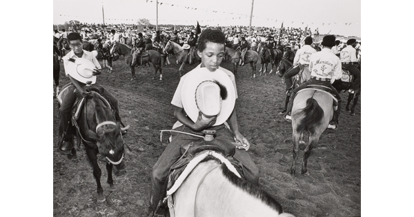 A black-and-white photograph of a large gathering of people on horses; two Black boys in the foreground holding their cowboy hats to their hearts.