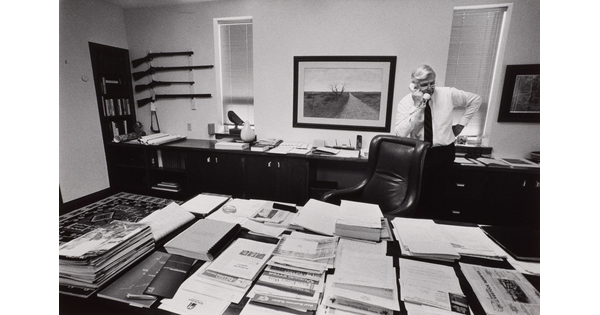 A black-and-white photograph of an older White man talking on a phone behind a large desk piled with papers.