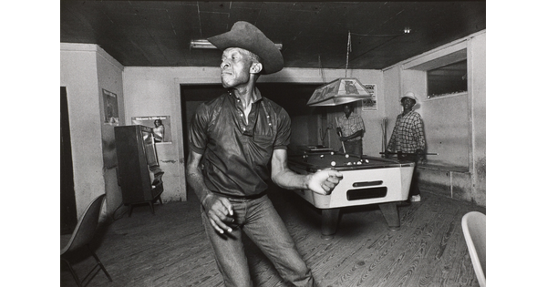 A black-and-white photograph of a Black man wearing a cowboy hat dancing in a pool hall in front of a billiards table.