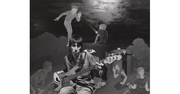 A black-and-white photograph of a young White man playing an electric bass guitar in front of a mural of people smoking and drinking.