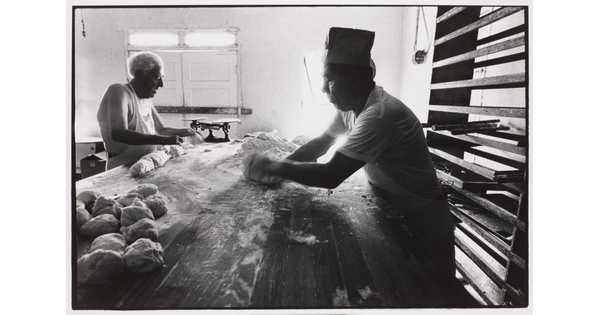 A black-and-white photograph of two men rolling dough on a long table covered in flour.