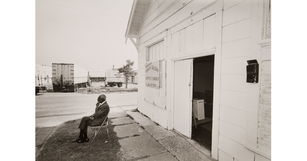 A black-and-white photograph of an older Black man in a suit sitting on a folding chair in front of a white building.