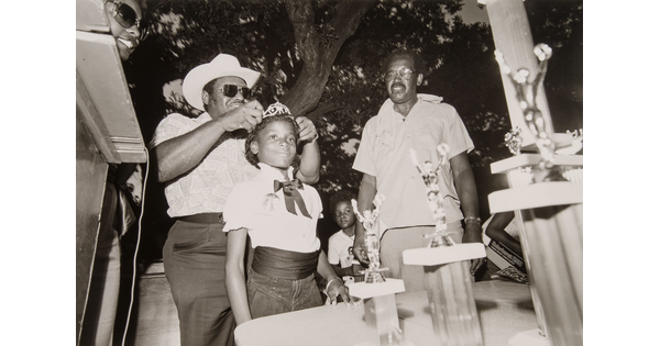 A black-and-white photograph of a young Black girl having a tiara placed on her head as others watch.