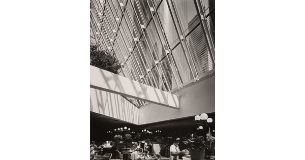 A black-and-white photograph of a cafeteria under a ceiling of windows that look up to a city skyline.
