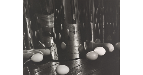 A black-and-white photograph of eggs on a wood surface reflected and distorted by an upright, wavy mirrored surface.