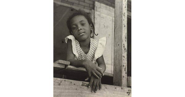A black-and-white photograph of a young Black girl in a gingham top leaning out of a window.
