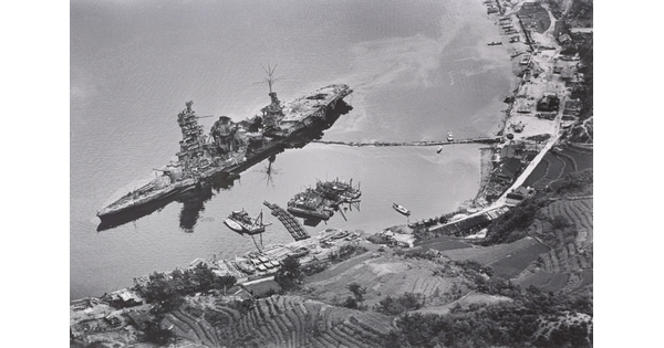 A black-and-white photograph of an aerial view of a large, damaged ship just off a hilly coast.