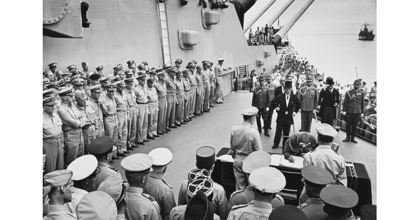 A black-and-white photograph of soldiers standing on the deck of a ship watching a smaller group of figures sign papers at a table.