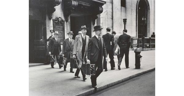 A black-and-white photograph of four White men in suits and hats exiting a city building and each holding a briefcase tethered to the other briefcases.