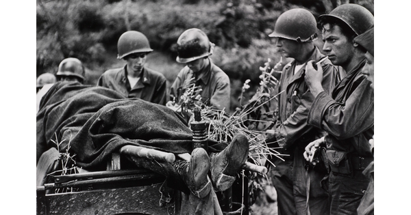 A black-and-white photograph of several helmeted White soldiers standing next a body covered in a blanket on a stretcher.
