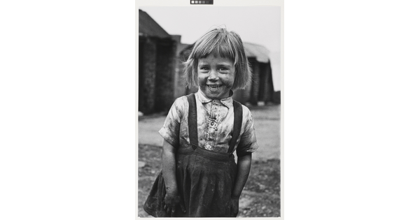 A black-and-white photograph of a smiling, coal-smudged young girl with fair skin and short hair, wearing in a dirty button-down shirt and skirt.
