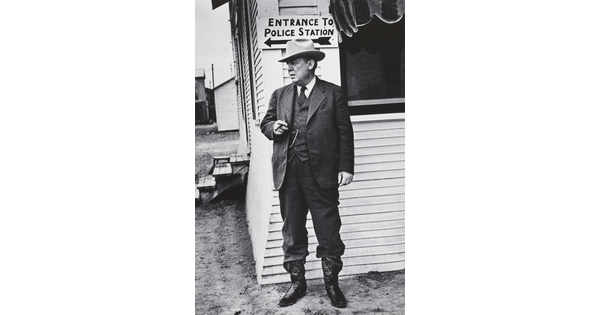 A black-and-white photograph of a White man in a three-piece suit, bowler hat, and cowboy boots, smoking a cigar and standing next to a clapboard building.